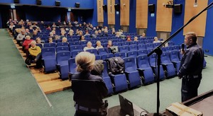 zdjęcie wykonane zza pleców policjanta i policjantki. Przed nimi widownia sali konferencyjnej z seniorami.